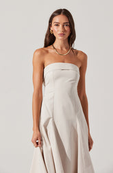 A woman is wearing a strapless white dress with a fitted bodice and pleated skirt. She accessorizes with gold jewelry, including a chain necklace and large earrings.